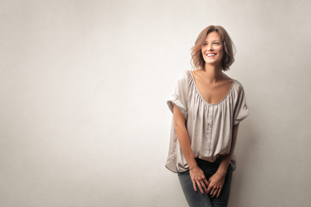 Woman wearing dark jeans and beige blouse smiling leaning against beige wall