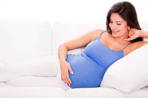 Pregnant woman wearing bright blue tank laying sideways on couch looking at belly