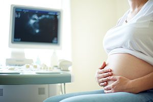 Woman from the neck down sitting in front of ultrasound holding pregnant belly